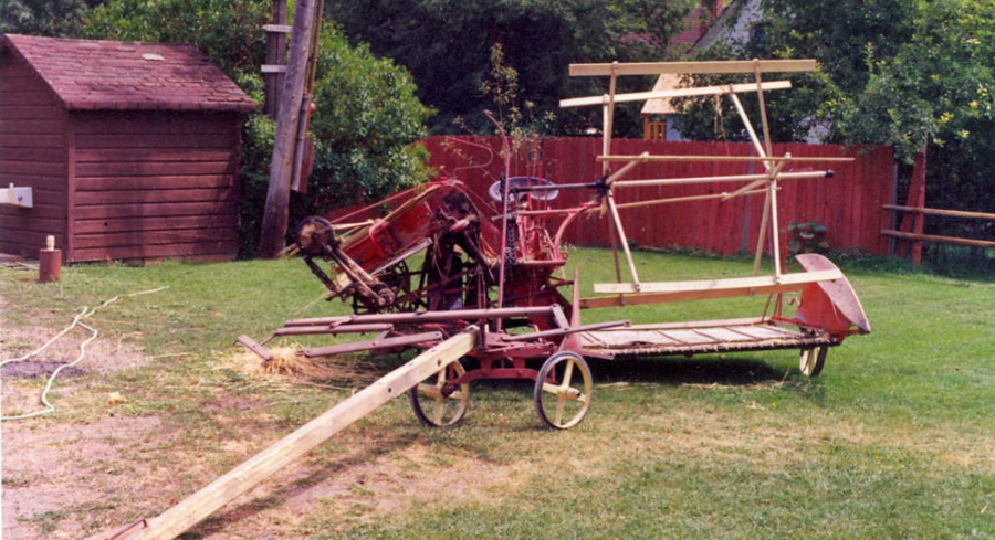 An old wooden farm machine with wheels stands on grass next to a small wooden shed. There are trees and a red wooden fence in the background. A pole and some ropes are seen lying on the ground.