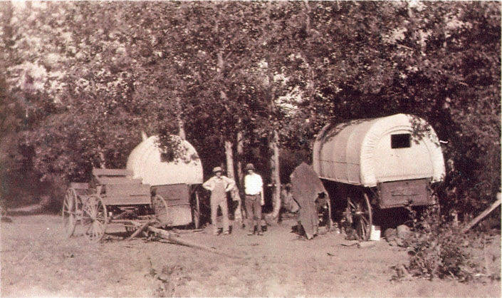 Two covered wagons are positioned among trees. Three people stand nearby, with one appearing to be leaning against the wagon on the left. The area is grassy with some branches scattered on the ground.