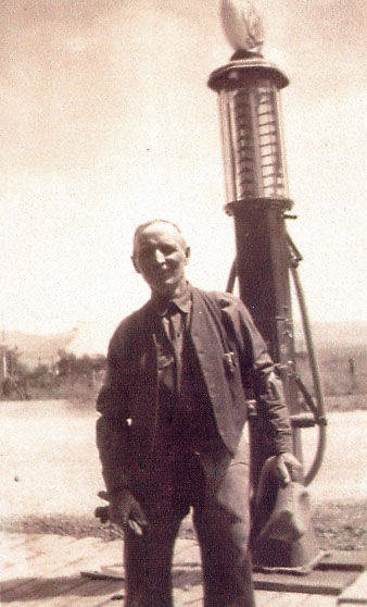 A man wearing a suit stands in front of an old-style gas pump, holding a hat in one hand. The background is an open area with distant trees or vegetation.