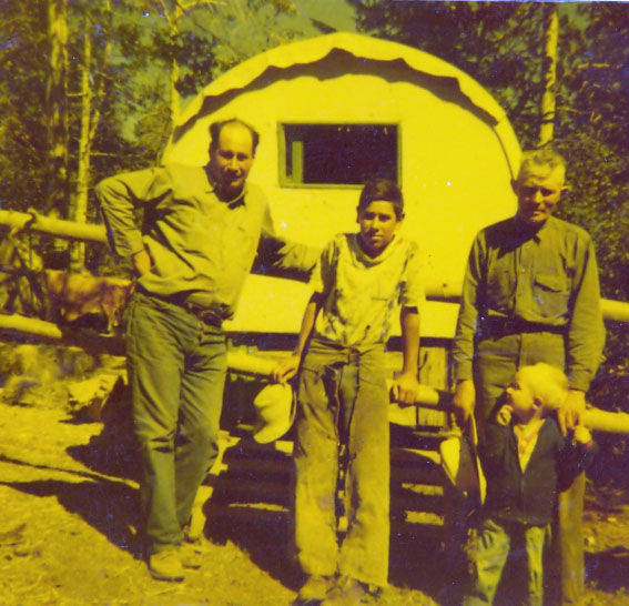 Several people stand in front of a covered wagon in an outdoor setting. One adult holds a hat, while another holds a small child. Trees and wooden fencing are visible in the background.