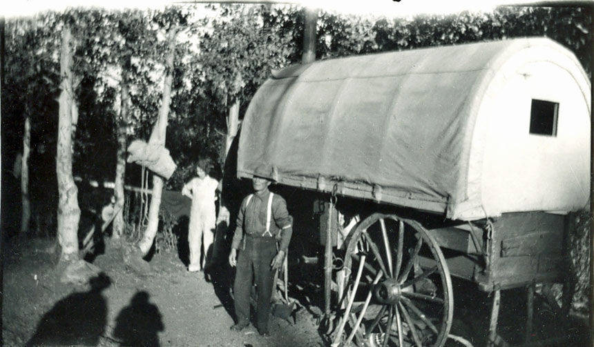 A group of people is gathered around a covered wagon with large wooden wheels in a wooded area. Some individuals are standing near the wagon, while others are partially hidden among the trees. The scene suggests an outdoor, rustic setting.
