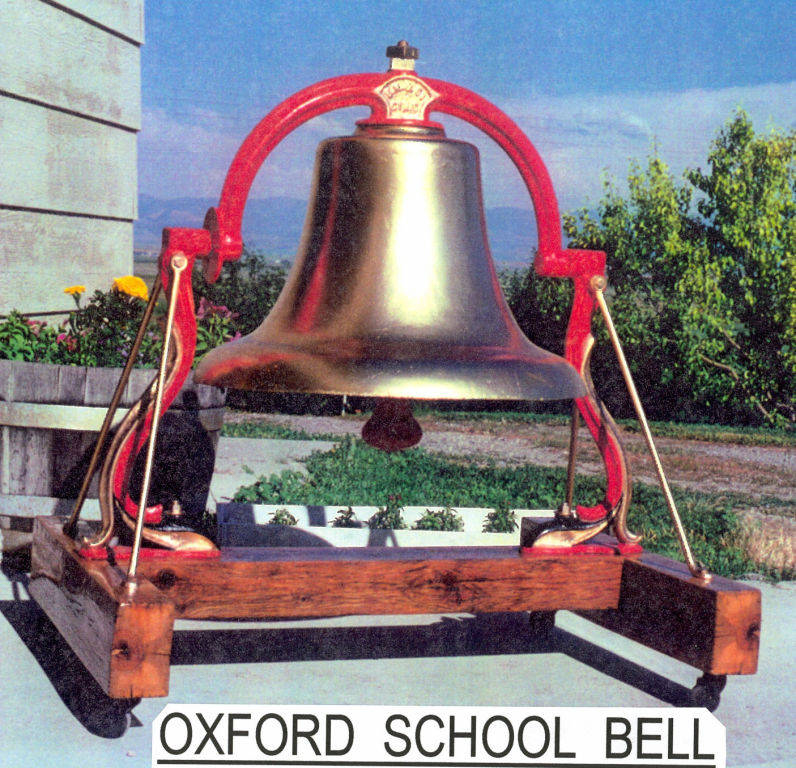 A large school bell mounted on a wooden frame with red brackets is placed outside. The background shows a garden with plants and a blue sky. Below the bell, there is text that reads "OXFORD SCHOOL BELL."