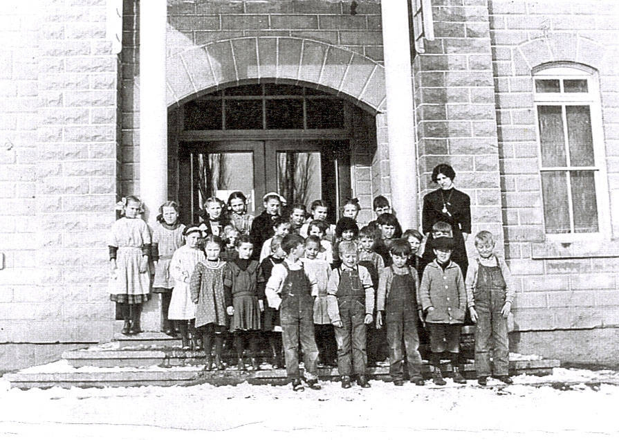 A group of children and a woman stand on the steps of a stone building with large columns. The children are dressed in a variety of clothing including dresses and overalls. The woman is wearing a long, dark dress. Snow is visible on the ground.