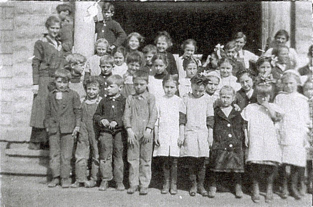 A group of children and a few adults standing together on the steps of a building. The children are dressed in various outfits, some with bows in their hair. They are positioned in several rows, with some older girls standing in the back. The building has a brick exterior and a large doorway.