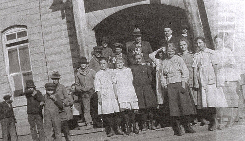A group of children and adults standing in front of a large building with an arched entrance. The children, mostly girls, are wearing dresses, while the adults in the background have hats and coats. Some boys are wearing caps and jackets. There are windows visible on the side of the building.