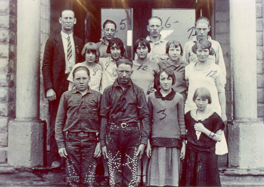 A group of thirteen people standing in front of a building with stone columns. The group consists of men, women, and children. Each person has a number written on them, ranging from 1 to 13. The individuals are arranged in three rows, with children at the front and adults at the back. No other visible text is present.