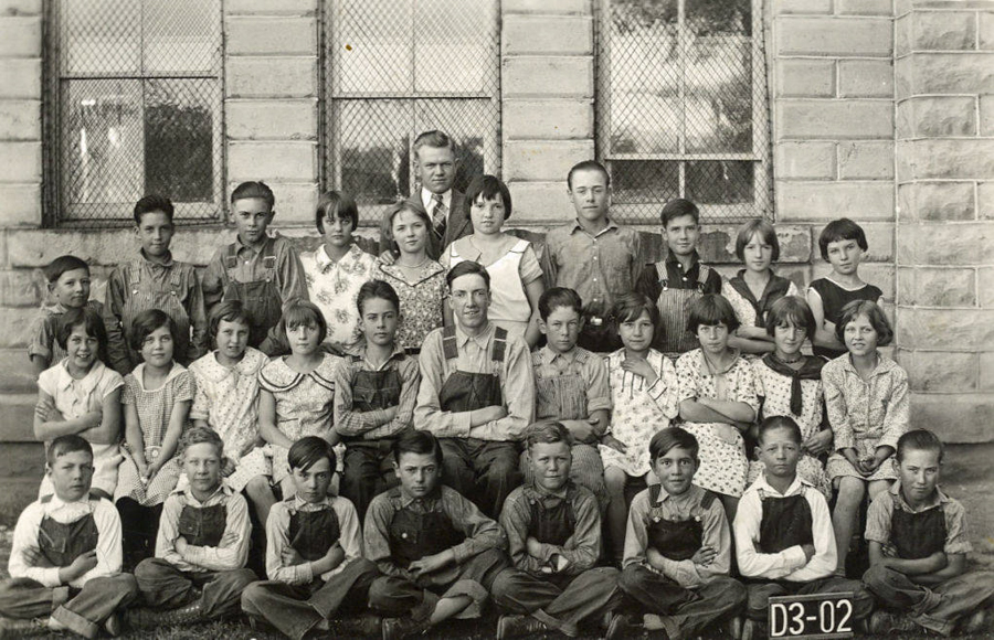 A group of children and two adults are posed in front of a stone building with wire-meshed windows. The children are arranged in three rows, with some seated and others standing. They are wearing a variety of clothing including dresses and overalls. A small sign in front reads "D3-02".