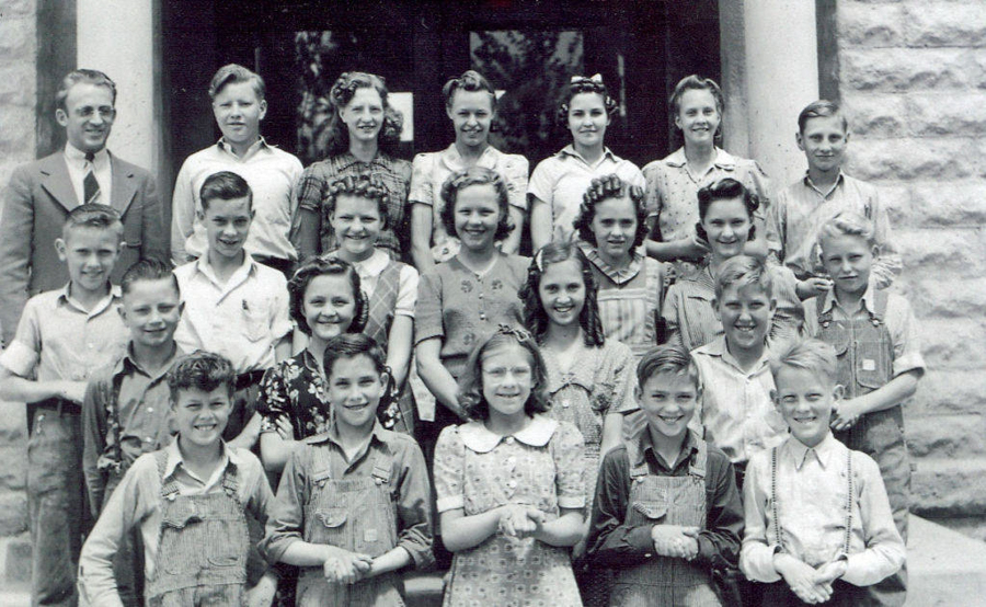 A group of children and an adult standing in front of a stone building. The group includes boys and girls, with the boys mostly wearing overalls or shirts, and the girls dressed in patterned dresses. The adult is wearing a suit and tie. The individuals are arranged in several rows, smiling at the camera.
