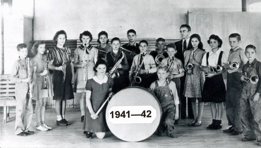 A group of young people is standing in a room, each holding a musical instrument such as clarinets, trumpets, and saxophones. At the front, a girl is kneeling beside a large drum that is labeled "1941—42." There are benches and a wooden structure in the background.
