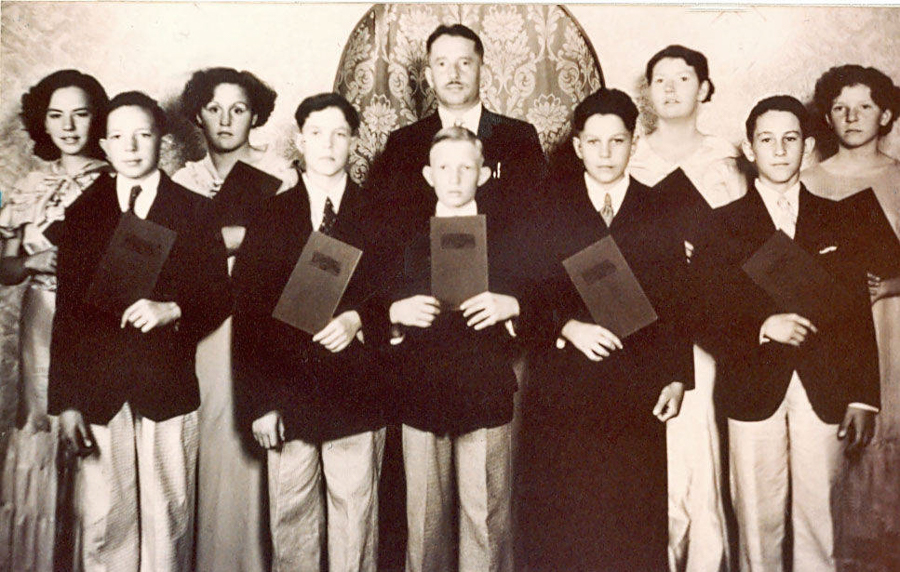 A group of ten people, consisting of six young men and four young women, standing in front of a patterned backdrop. The young men are wearing suits and ties, holding folders or diplomas. The young women are dressed in formal gowns. The central man, slightly older, is distinguished with a taller stance.