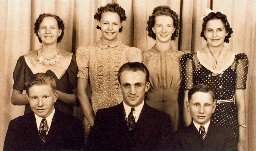 Three women stand in the back row, smiling, each wearing dresses with distinct patterns and collars. In front of them, two young men and one older man sit, all wearing suits and ties. The setting is indoors, and a curtain is in the background.