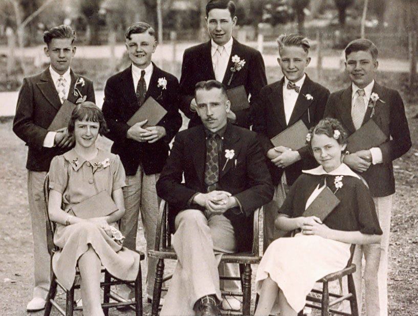 A group of eight people posing outdoors. Five young men and two young women stand behind an older seated man in the front row. The men and women are dressed formally, with flowers in their lapels. The young people hold diplomas or certificates. The setting is a park or garden area with trees in the background.