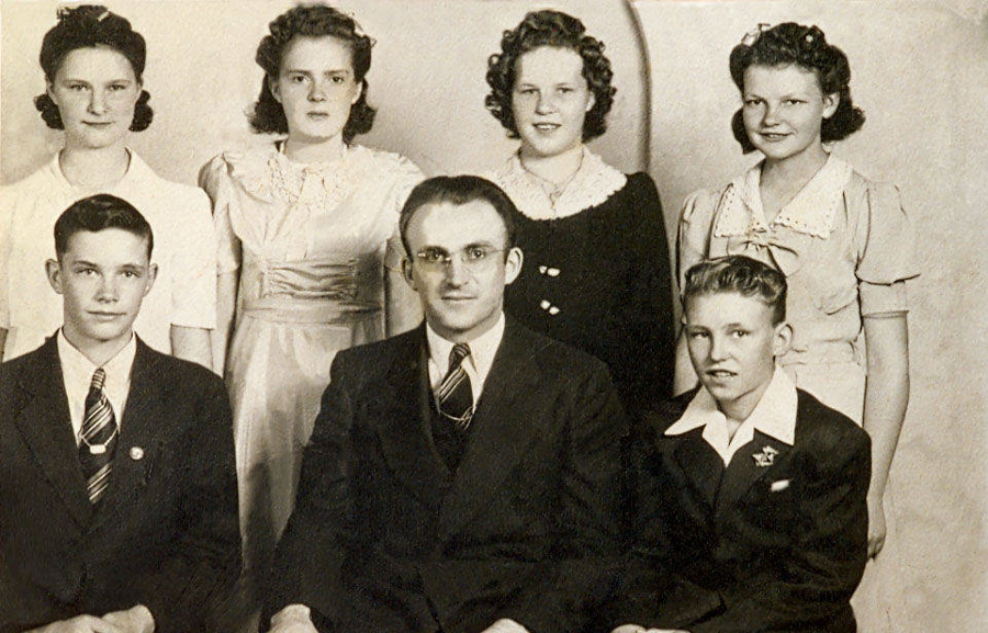 A group of six people, with four women standing in the back and two men seated in the front. The women are wearing dresses and have similar hairstyles. The men are dressed in suits, with ties. The setting suggests a formal or family portrait.