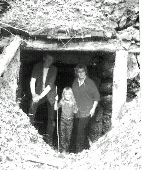 A group of three people standing inside the entrance of a cave or tunnel. The opening is reinforced with wooden beams, and the entrance is surrounded by rocks and earth. The group includes a young girl holding a stick, flanked by two adults. The area around them appears rugged and natural.
