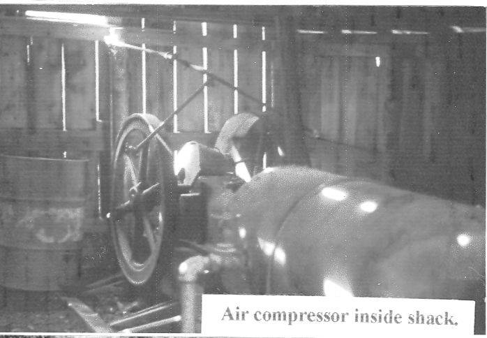 An air compressor inside a wooden shack, with a large wheel and pipes visible. There is a drum next to it and light coming through the gaps in the wooden walls. A label reads "Air compressor inside shack."