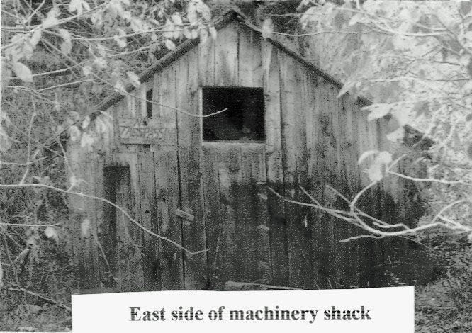 A wooden shack with a peaked roof surrounded by foliage. There is a square window opening near the top. The structure appears weathered. Text below the image reads, "East side of machinery shack."