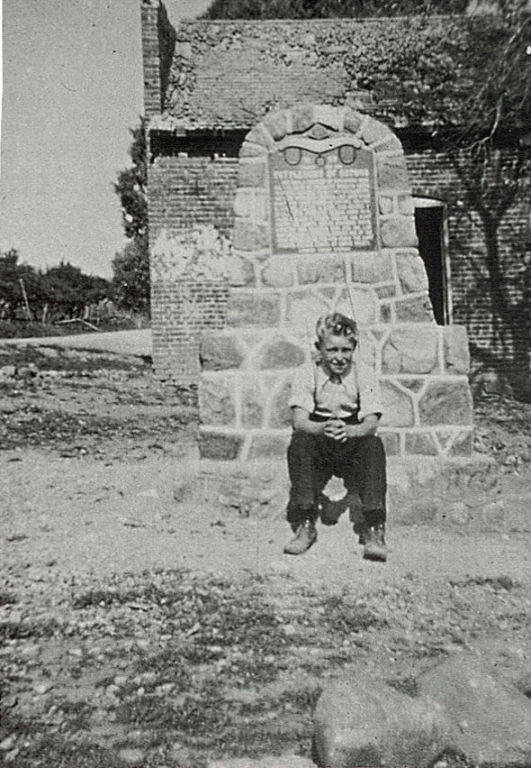 A child sits on a stone structure, which has a plaque with some text on it. The child is wearing a light-colored shirt and dark pants. Behind the structure, there is a brick building. Trees are visible in the background.
