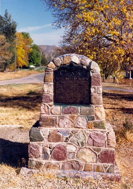 A stone monument with a plaque stands by a roadside in a grassy area. The plaque is titled "Settle Men of Dakota" and contains additional text, though it is not fully legible. In the background, there are trees with yellow leaves and a road leading into the distance.