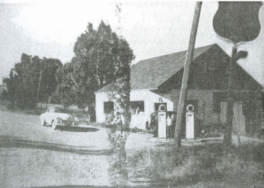 A rural scene with an old gas station featuring two visible gas pumps and a small building in the background. A car is parked to the left near the gas station, and there are some trees around the area. A large utility pole is in the foreground.