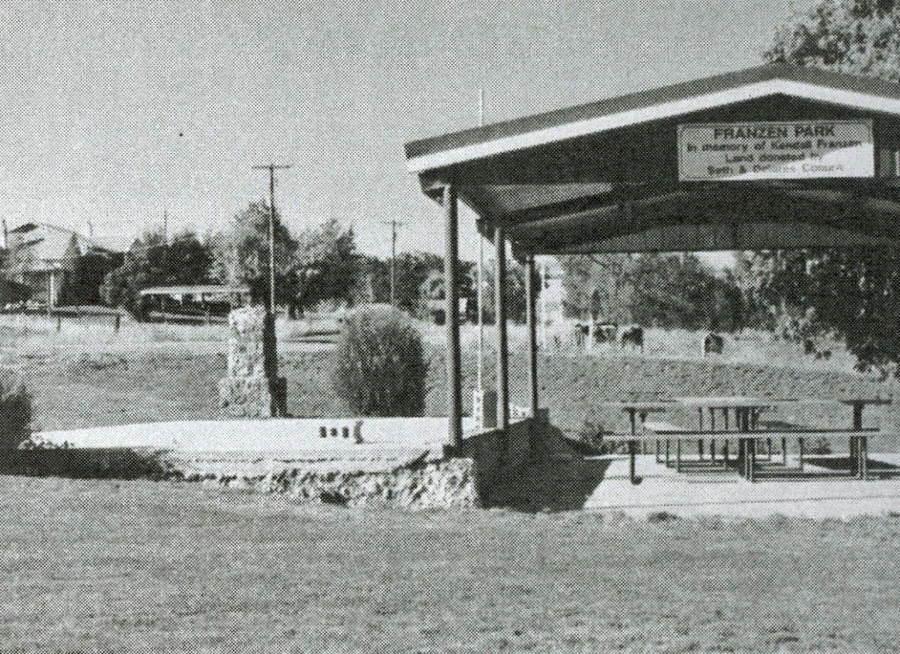 A covered area with picnic tables beneath it in a park setting. The sign reads "FRANZEN PARK in memory of Richard Franzen and descendants. Don & Catherine Coman." There are trees and utility poles in the background.