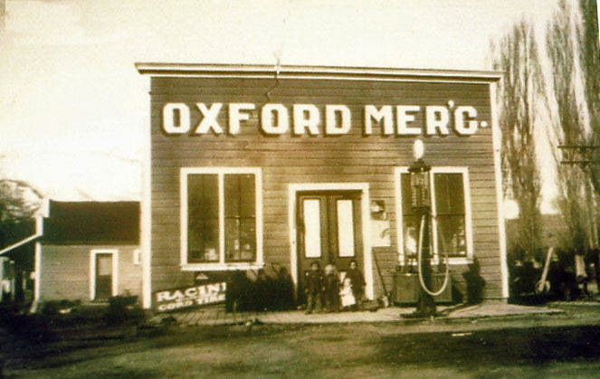 A wooden building labeled "Oxford Merc." The facade has two windows and a central door. A gas pump stands to the right of the entrance. Several people, including children, are gathered near the door. A sign to the left of the building reads "Racine Country Time." Trees and a small house are visible in the background.