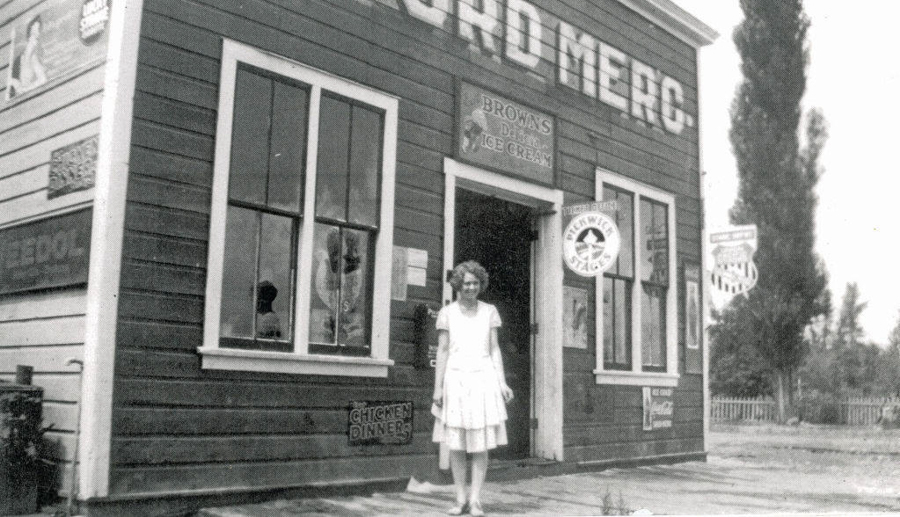 A woman stands in front of a wooden building with large windows and a door. The signage on the building includes "BROWN'S ICE CREAM," "CHICKEN DINNERS," and "BUY WHEAT FLAKES." Above the door, the words "RD MERC" are visible, along with various other smaller signs. Trees and a fence can be seen in the background.