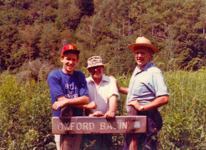 Three men standing behind a wooden sign that reads "Oxford Basin" in a lush, green forest setting. One man on the left wears a red cap and a shirt with partially visible text. The man in the middle is wearing a light-colored hat and a white shirt, while the man on the right is wearing a straw hat and a light blue shirt.