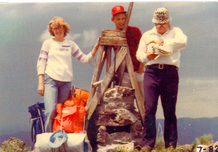 Three people standing outdoors near a wooden and stone structure. One person on the left is wearing a white sweatshirt with purple stripes and "Bergen" text, jeans, and is resting a hand on the structure. A person in the middle is wearing a red hat with an "H" on it and a red shirt. The person on the right is wearing a white shirt, dark pants, and a hat, while looking at papers. There are backpacks on the ground and hills in the background. The date "7-82" is in the lower right corner.