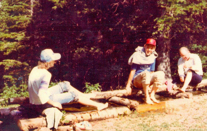 Three people are sitting on a log structure in a wooded area. The first person wears a light-colored cap and has their feet in water. The second person, wearing a red cap and with a sweatshirt draped over their shoulders, is also sitting with their feet in water. The third person is seated slightly apart, looking down at something in their hands. The background is filled with trees and greenery.