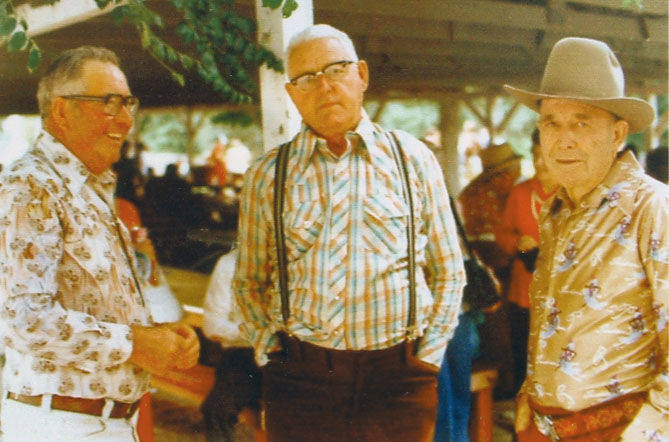 Three men standing together in a casual outdoor setting. The man on the left wears glasses and a patterned shirt, the man in the middle wears glasses, a plaid shirt, and suspenders, and the man on the right wears a cowboy hat and a patterned shirt. There are people and table benches in the background.