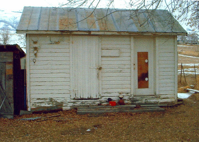A small, weathered white wooden shed with a metal roof. The door is centrally located, with a makeshift lock. To the right of the door, there is a window covered with a board that has two circular cutouts. Near the base of the shed, there are several items including a red container and some scattered wooden planks. Bare tree branches hang overhead, and patches of snow are visible on the ground around the shed.