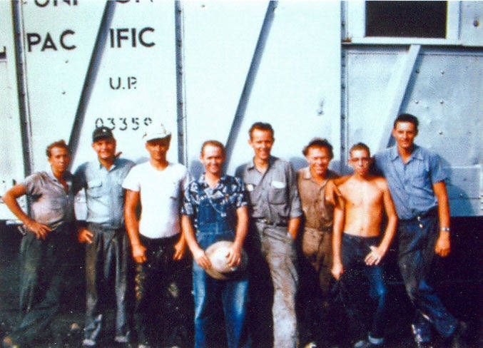 Eight men standing in front of a train car with the words "PACIFIC" and "U.P. 03350" visible. One man in the center is holding a ball. Some men are wearing work clothes, while others are in casual attire.