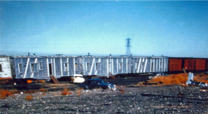 A freight train with several boxcars in a rail yard. There are two cars parked near the train, and a utility tower can be seen in the distance. The ground is covered in gravel and some scattered plants.