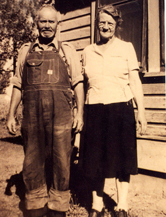 An older man and woman are standing together outdoors. The man is wearing overalls and a shirt, while the woman is dressed in a blouse and skirt. They are posed in front of a wooden building, possibly a house, with a window visible behind them. There are trees in the background.