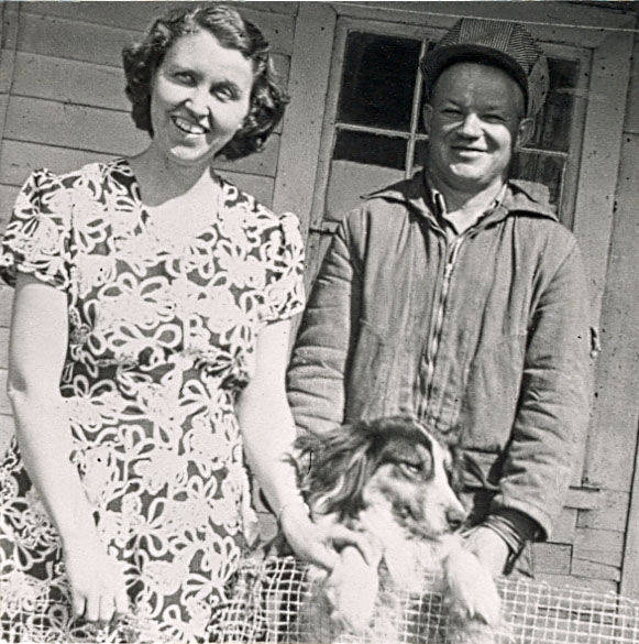 A woman in a patterned dress stands next to a man wearing a jacket and cap. They both smile while holding onto a dog in front of a wooden house with a window and door.
