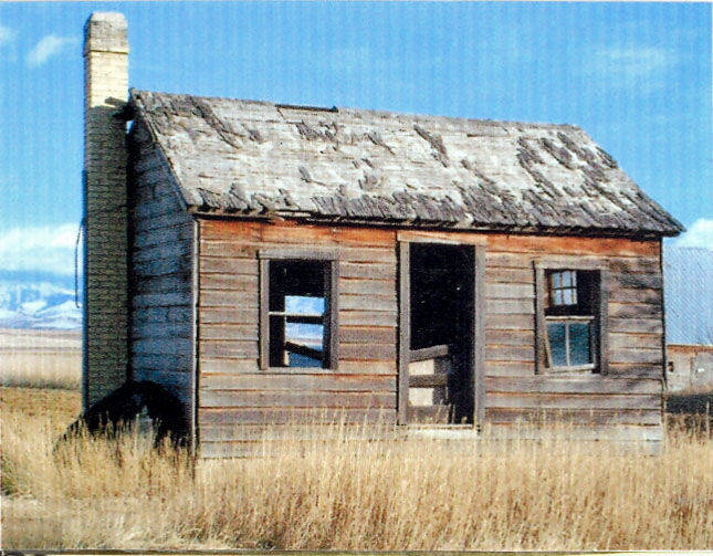 An old, weathered wooden cabin with a tall brick chimney. The roof appears worn, and there are two windows with some missing panes. The entrance has no door, revealing the interior. The cabin is situated in a grassy open field with mountains in the background.