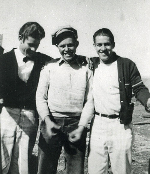 Three young men standing outdoors, smiling. They are wearing casual clothes, with two of them in sweaters and one in a cardigan. Two of the men have their arms around the one in the center. There is no visible text.