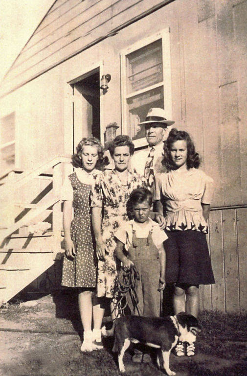 A group of five people stands outside near a wooden building with stairs leading to a door. Three women, one man, and one child are part of the group. The women and the child are in front, with the man slightly behind them. There is a dog with a leash in front of the group, standing on the grass.