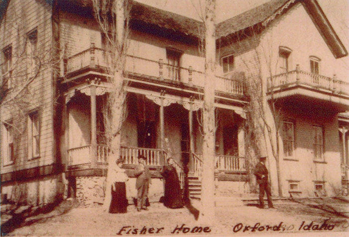A group of five people stands in front of a large two-story house with a porch and balcony. The trees around the house have no leaves. The words "Fisher Home Orford Idaho" are written below the scene.