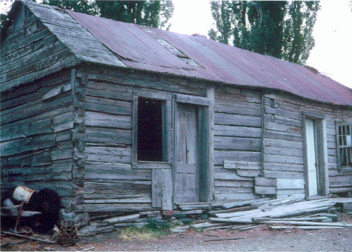 A weathered wooden cabin with a metal roof. The structure has a slightly open door, a window without glass, and another window with some panes missing. Pieces of wood are scattered around the ground near the building. In the foreground, there is a donkey with a saddle bending down near the cabin. Trees are visible in the background.