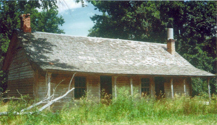 A single-story wooden house with a shingled roof, featuring two chimneys. The house has a porch supported by multiple columns. There are tall grasses and some trees surrounding the structure, with a branch leaning against the house on the left side.