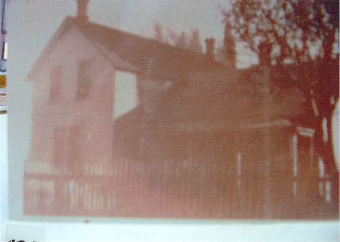 A house with a pitched roof and chimney, surrounded by a wooden fence. There are trees visible in the background.