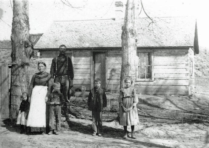 A family of five stands in front of a small wooden house. The group includes two adults and three children. The children are positioned on either side of the adults, and two large trees are also visible in the foreground. The house has a sloped roof and a single window.