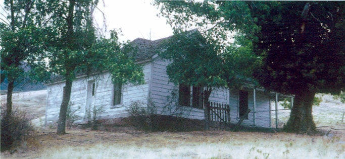 A small, weathered wooden house with a front porch sits amid a grassy area. There are several trees surrounding the house, and the area appears rural and overgrown. The house has a few windows, and a path or dirt road seems to lead up to it. The setting is quiet and secluded.