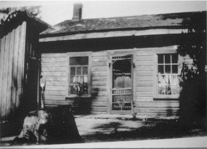 A small wooden house with a sloped roof and two windows on either side of a central door. The windows have curtains inside. A tree stump is visible in the foreground to the left, and there is a wooden annex to the left of the main house. Shadows of foliage appear on the right side of the structure.