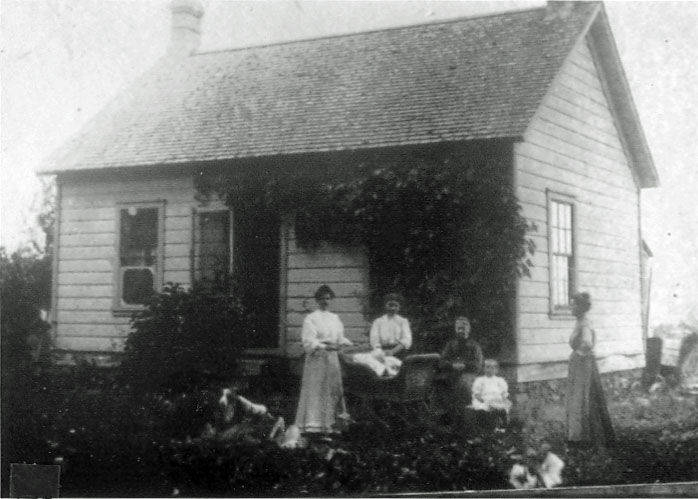 A group of people, including women and children, are gathered in front of a small wooden house with a pitched roof. The house has two visible windows and is partially covered with foliage. A few dogs are present in the foreground. The people are dressed in period clothing.