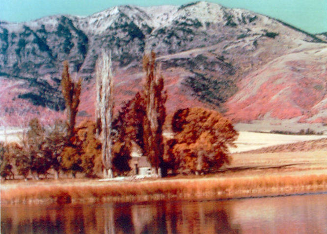 A serene landscape featuring a small house nestled among tall trees and autumn foliage. In the background, snow-capped mountains rise against a clear sky. A calm body of water reflects the scene in the foreground.