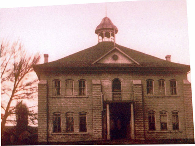 A large two-story building with a prominent central entrance and multiple windows. The structure features a small cupola on the roof. Tall trees are visible to the left and right sides of the building.
