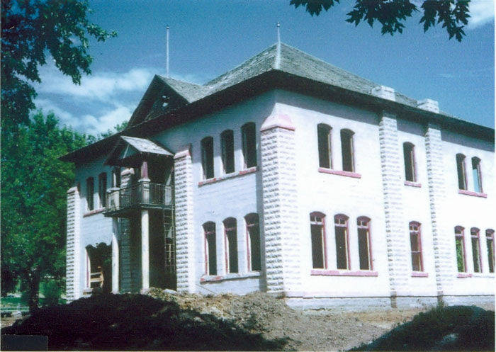 A two-story, rectangular building with a sloped roof and numerous windows. The walls are made of light-colored material with decorative trim around the windows and edges. There is a small balcony above the entrance, with a set of stairs leading up to it. The building is surrounded by a clearing, and trees are visible in the background.