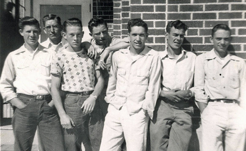 Seven young men stand together in front of a brick wall. Some have their arms around each other's shoulders. They are casually dressed in shirts and pants. One of them wears a patterned shirt, while the others wear plain clothing. The group appears relaxed and smiling.